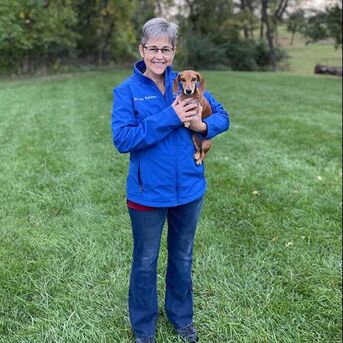 Dr. Jan Tallman, Owner & Veterinarian at Eastview Animal Hospital standing and smiling while holding her little dachshund dog 